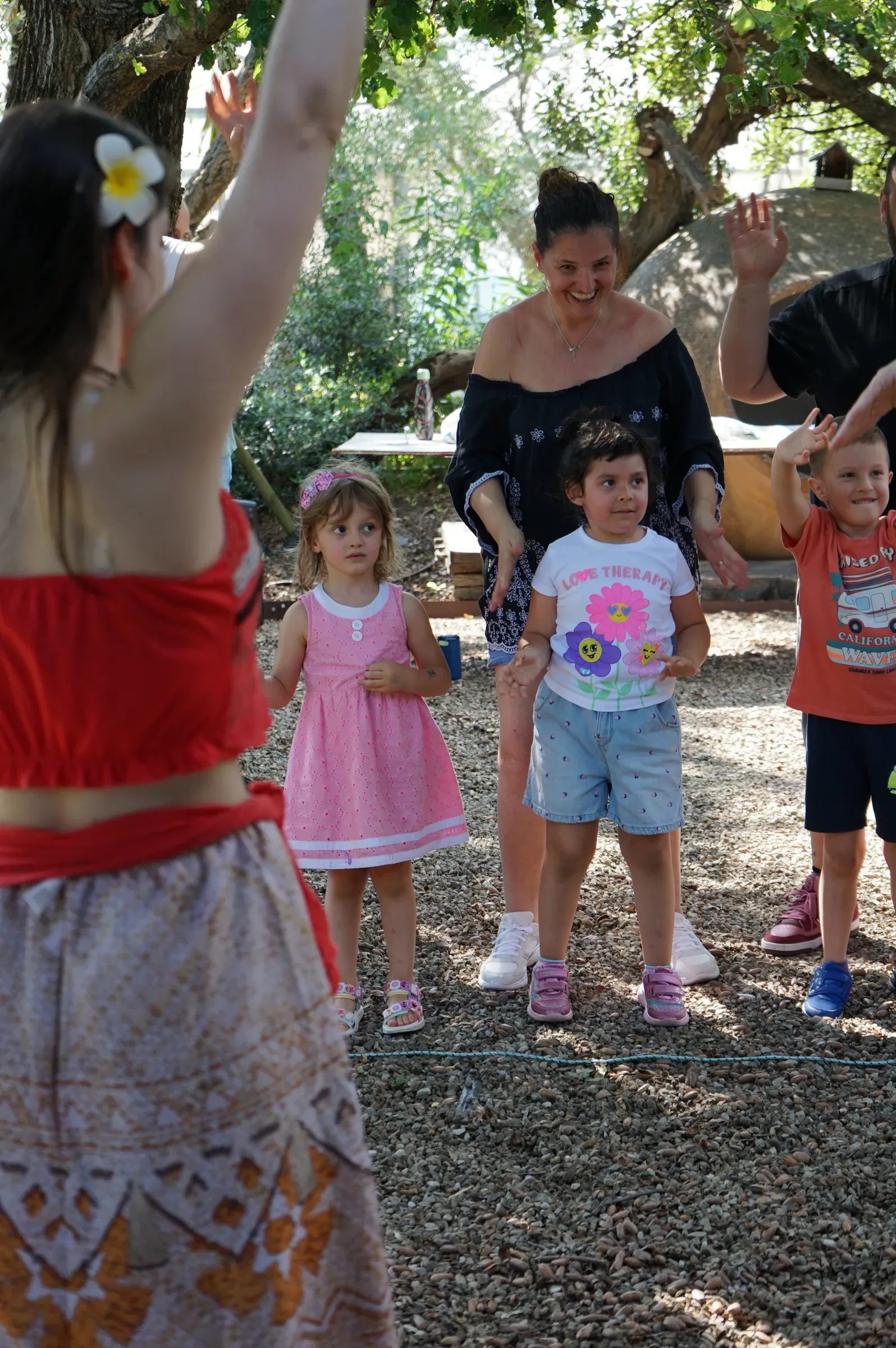 Bambini e genitori ballano sotto gli alberi, alcuni a braccia alzate, con Vaiana di spalle in primo piano