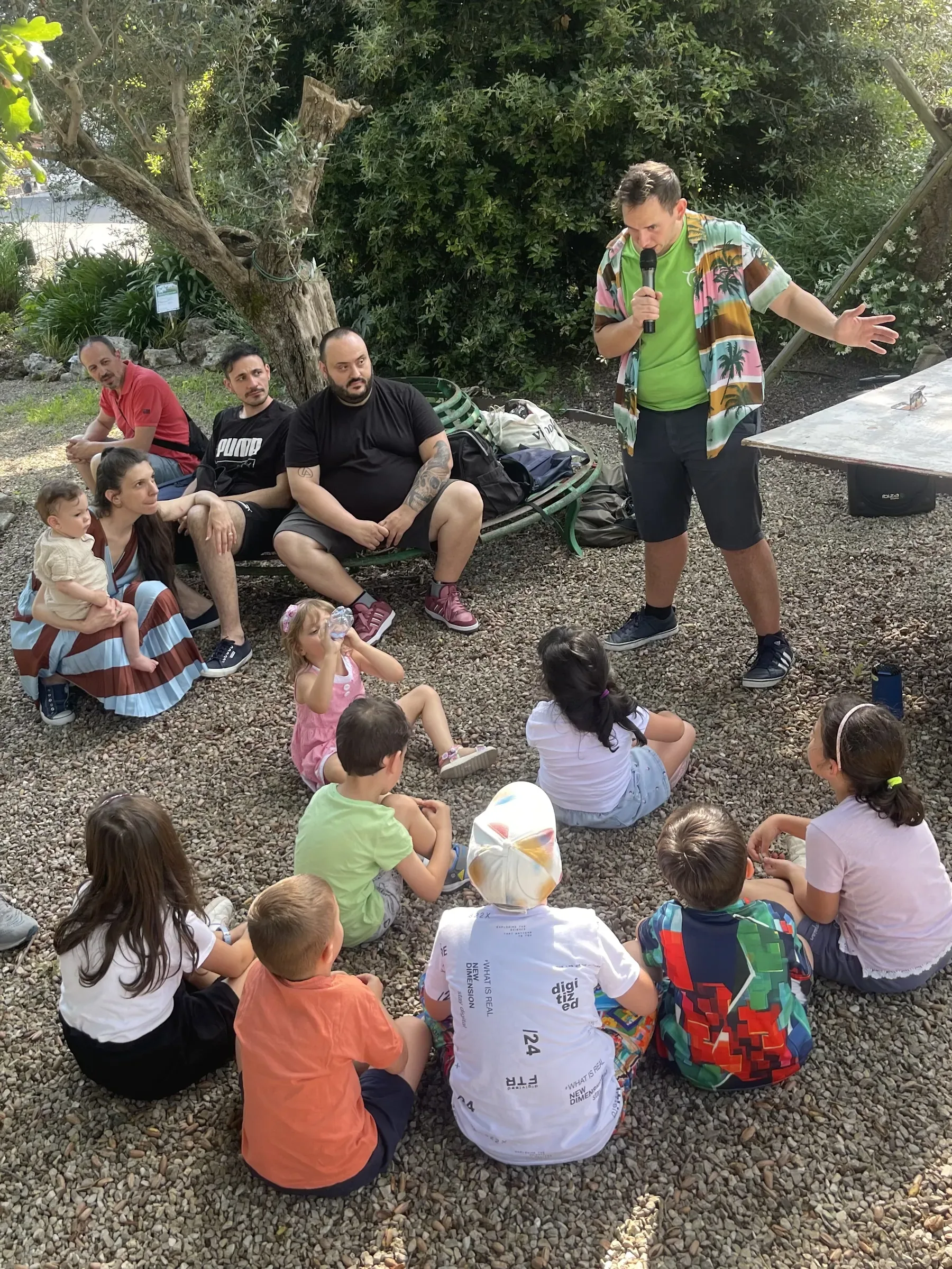 Un animatore in maglietta verde con il microfono parla ai bambini seduti sulla ghiaia, con alcuni adulti seduti dietro