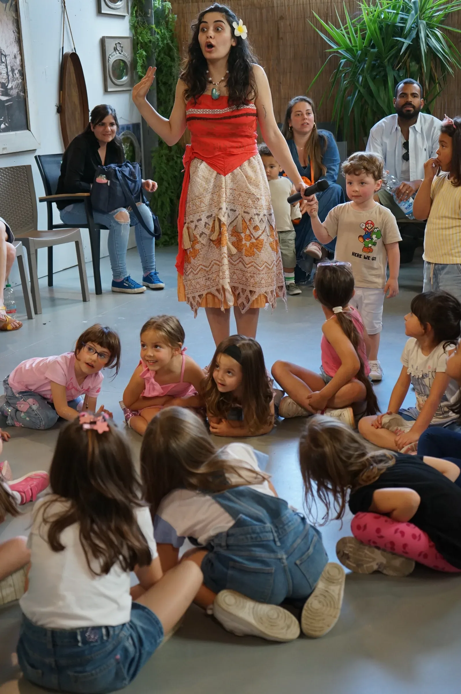 Vaiana in piedi, con il microfono in mano, parla ai bambini seduti in cerchio sul pavimento, con alcuni adulti seduti dietro