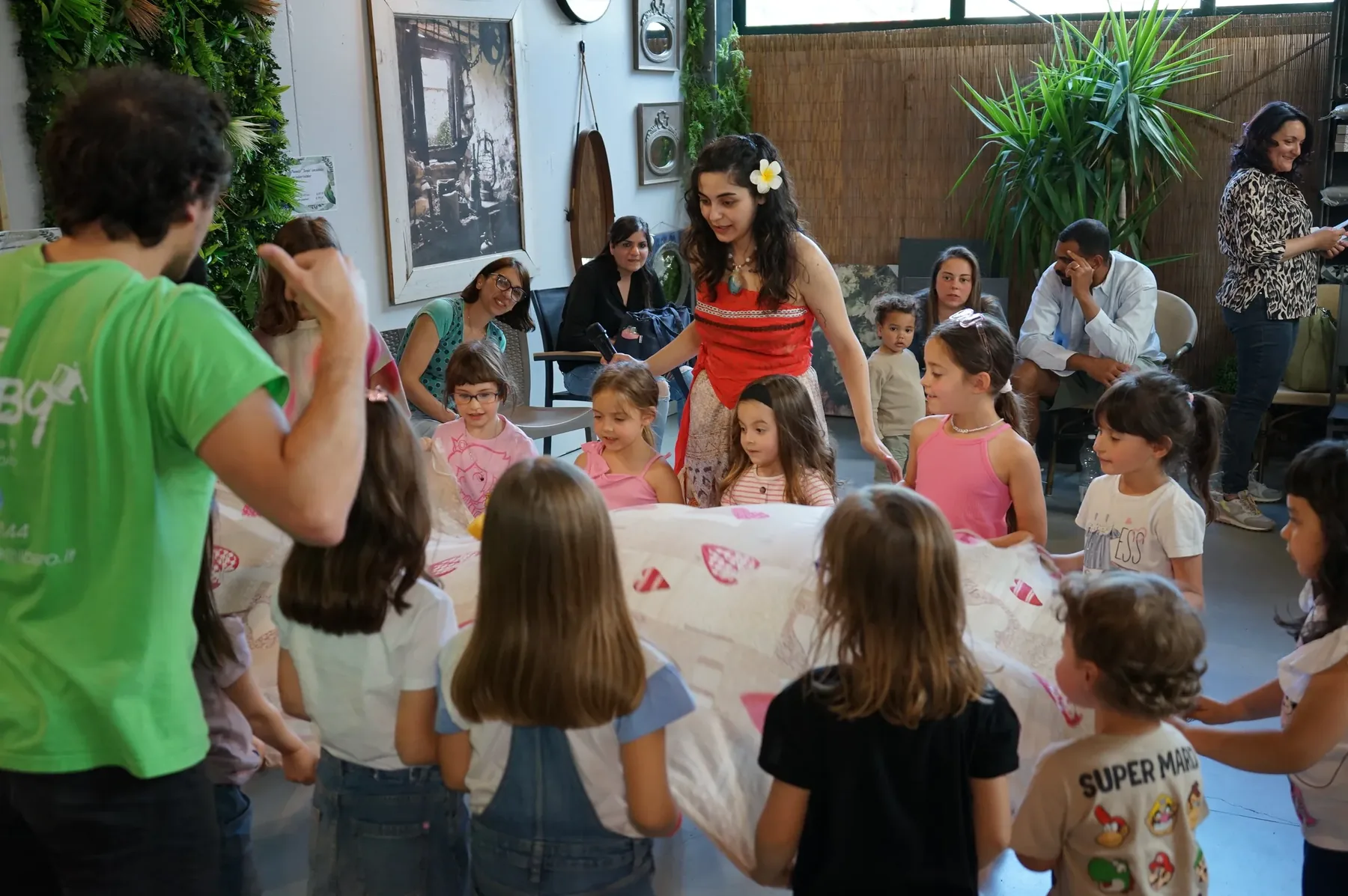 Un animatore in maglietta verde di spalle e i bambini in cerchio attorno al telo, con Vaiana in mezzo a loro e i genitori seduti sul fondo