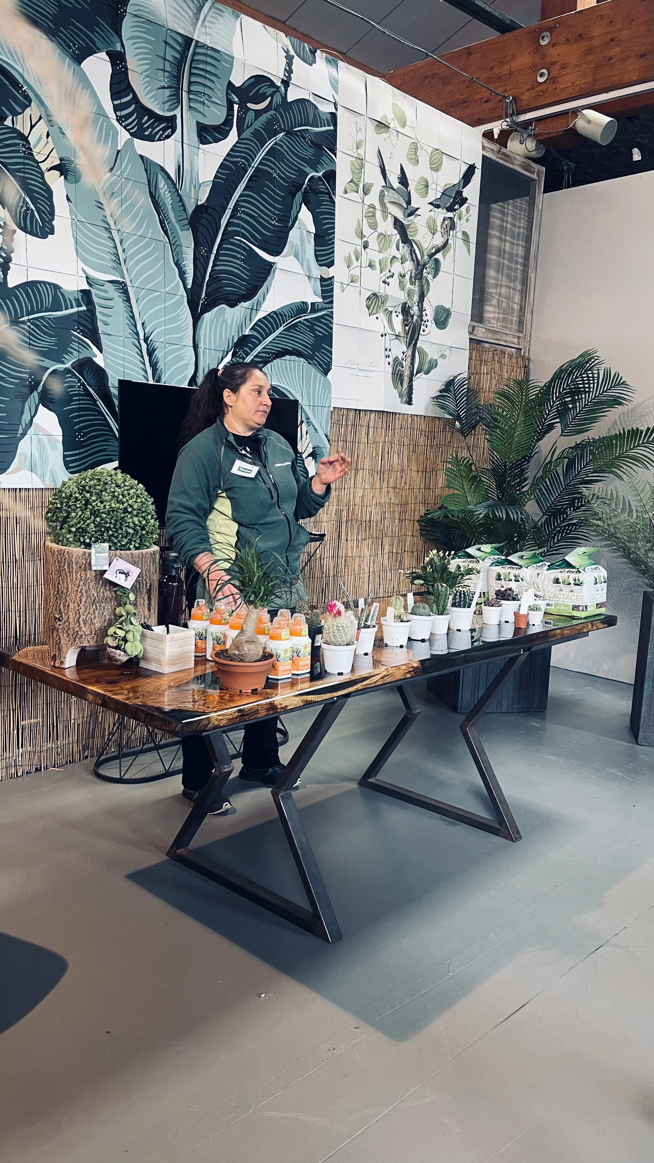 Una donna in giacca verde parla gesticolando dietro il tavolo lungo con i cactus, i flaconi e i sacchetti di terriccio