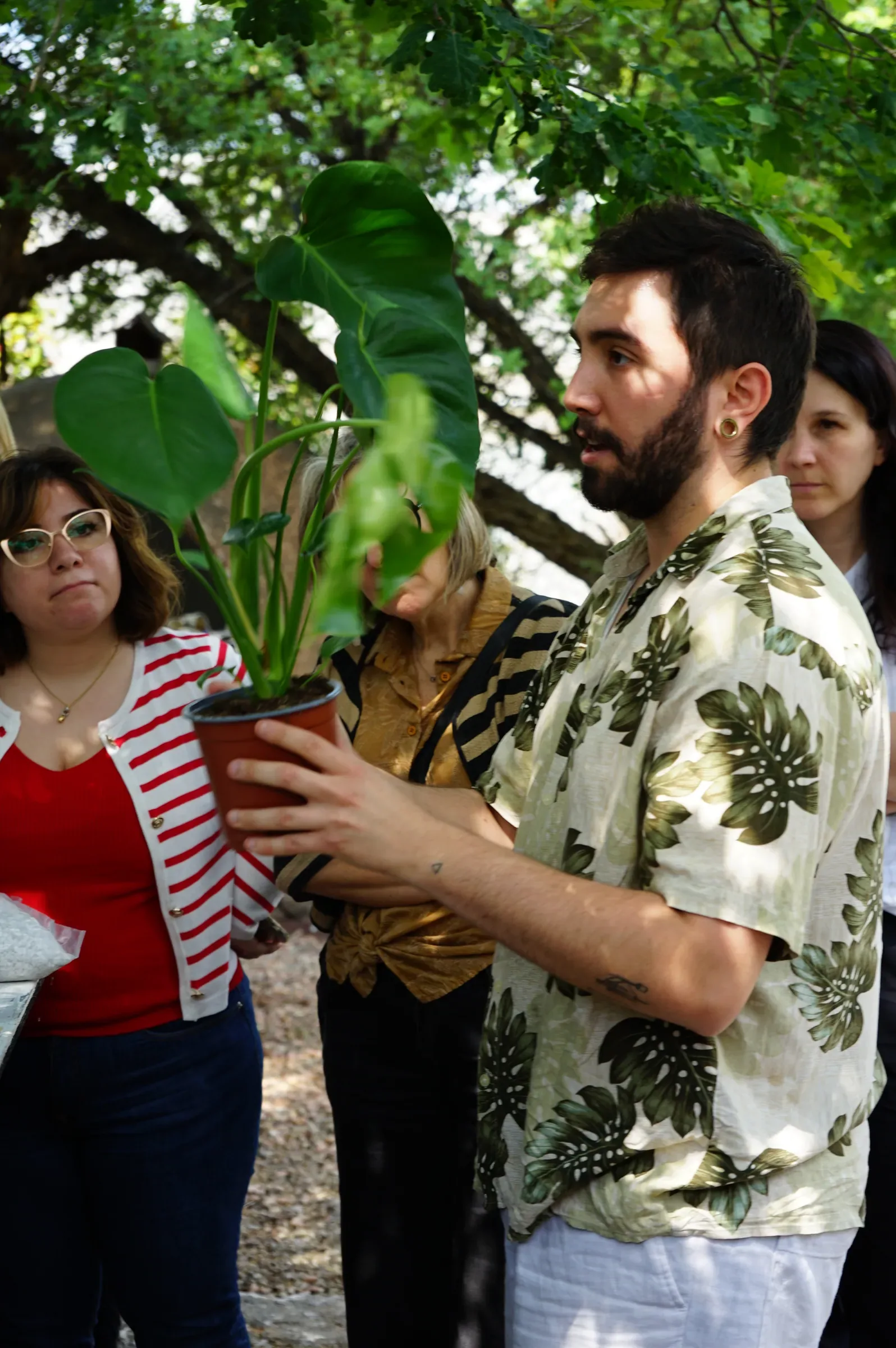 Un ragazzo con la camicia a foglie tiene una monstera in un vaso color terracotta, fra le partecipanti in piedi sotto un albero