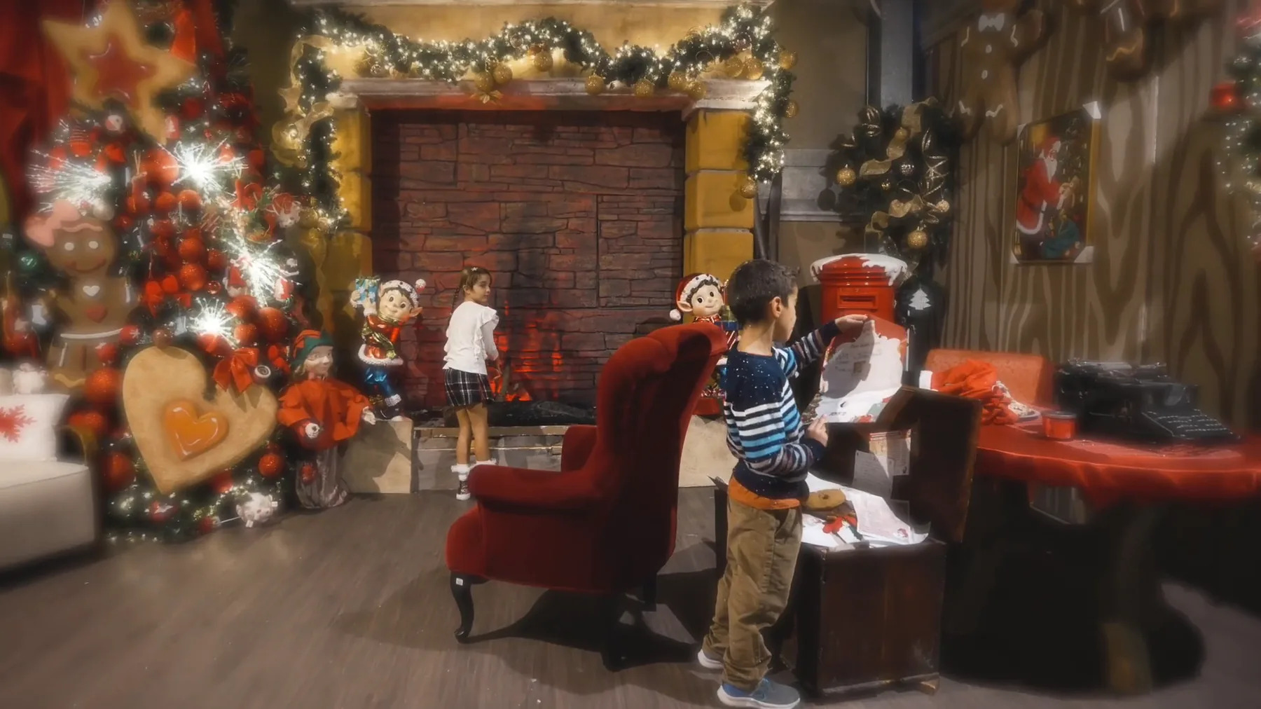 Nella casa di Babbo Natale un bambino tiene una lettera sopra un baule pieno di letterine, davanti alla cassetta rossa, fra la poltrona di velluto, il camino e l'albero addobbato