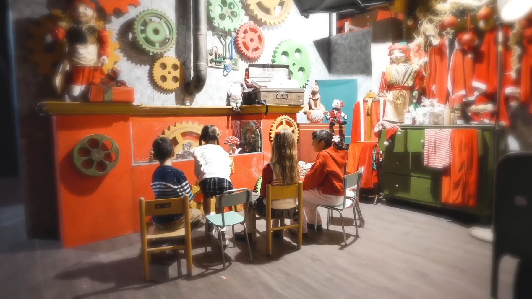 Bambini di spalle seduti sulle seggioline davanti al banco arancione del laboratorio di Babbo Natale, sotto la parete di ingranaggi colorati
