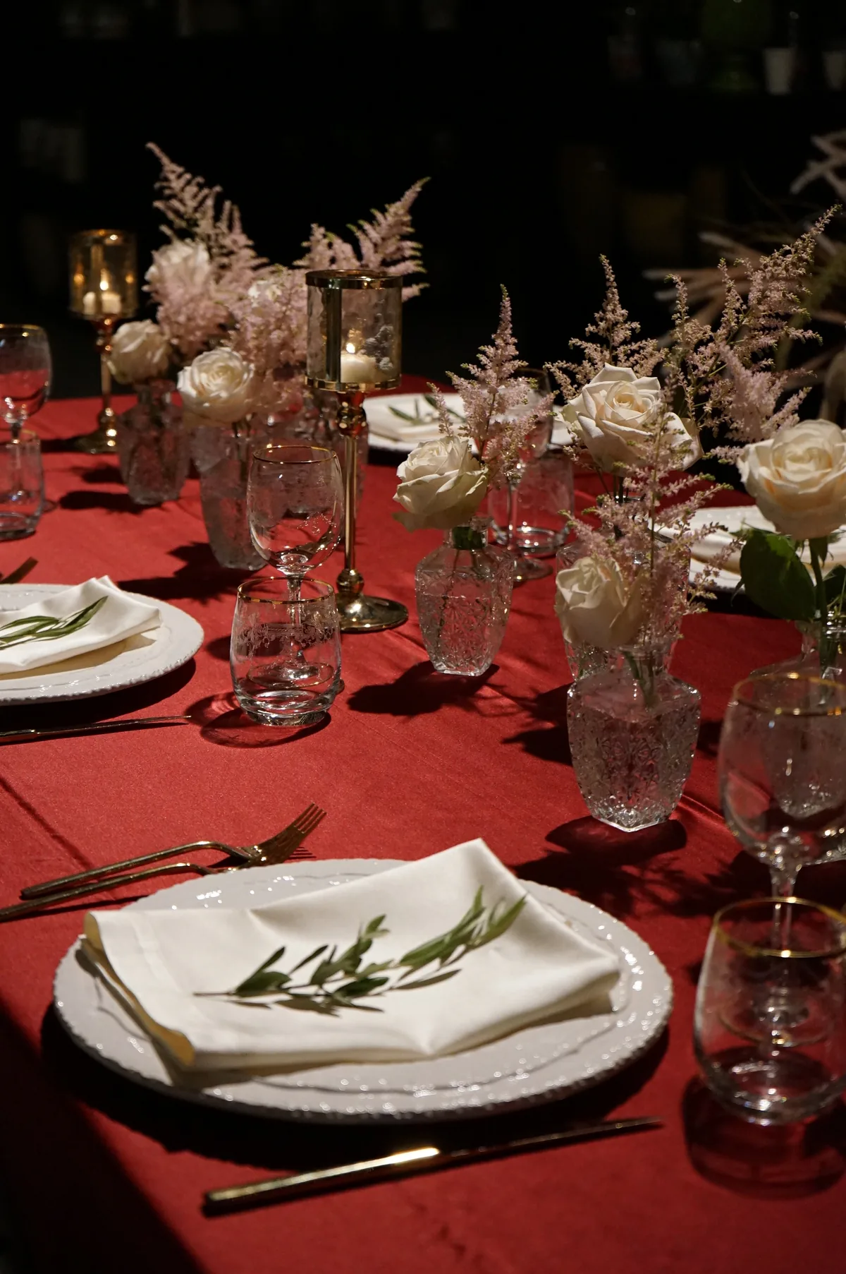 Coperti con tovaglioli bianchi e rametti d'ulivo sulla tovaglia rossa, con rose bianche e pennacchi rosa in vasetti di vetro lavorato