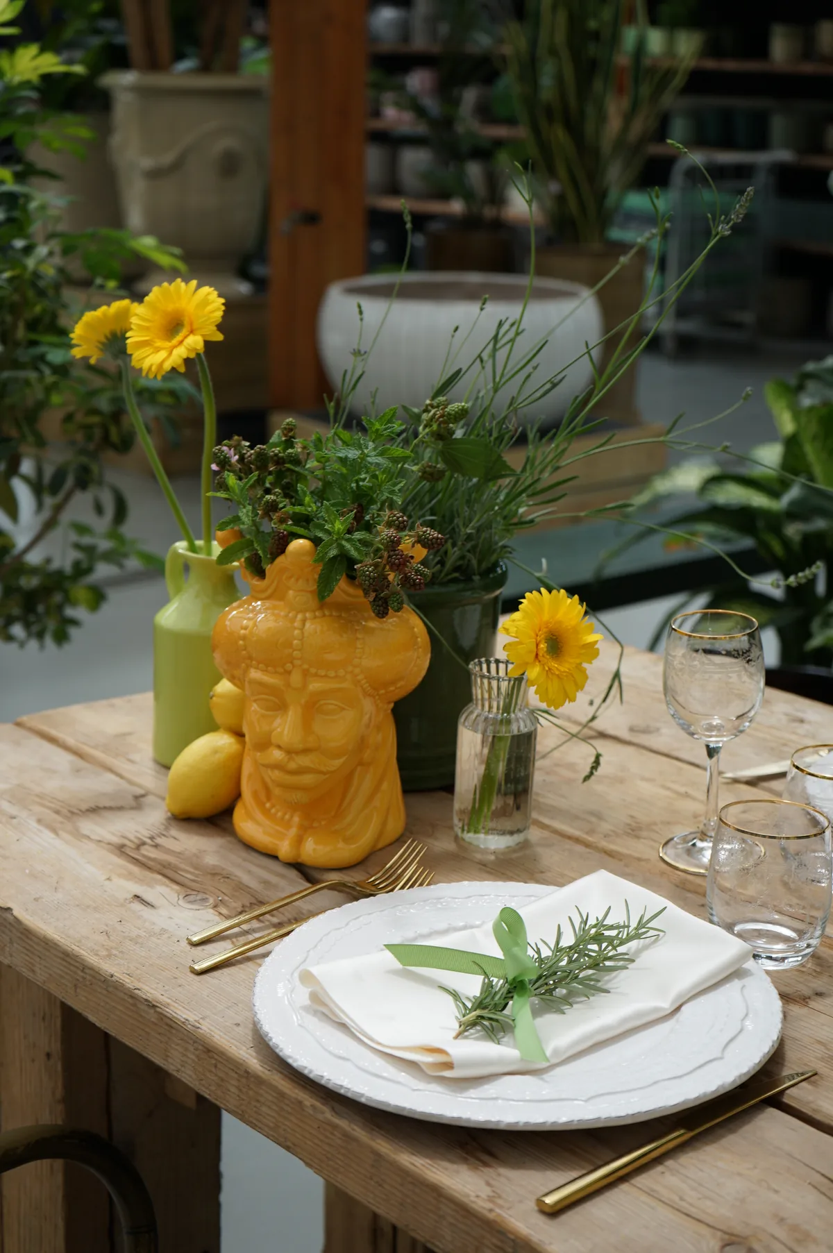 Vaso giallo a testa di moro con erbe e more, limoni, vasetti verdi e gerbere gialle su un tavolo di legno grezzo, davanti a un piatto con il tovagliolo e un rametto di rosmarino